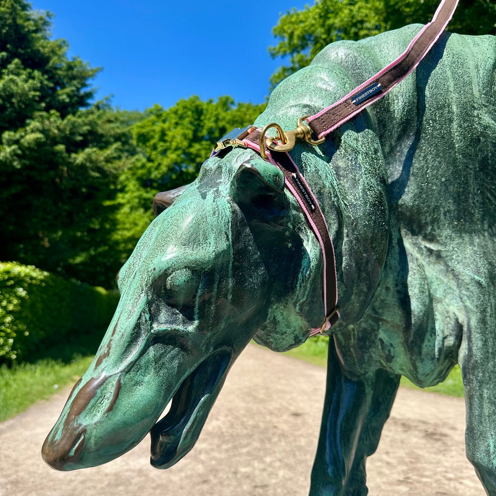 Hund trägt FINESTBOY-Bundle aus Halsband und Leine, 25mm, rosafarbenes Gurtband mit dunkelbraunem Canvas-Streifen und Vollmessing-Applikationen, vermessingter Metallsteckschnalle und Vollmessing-Karabiner, im Hamburger Stadtpark an einer Hundeskulptur