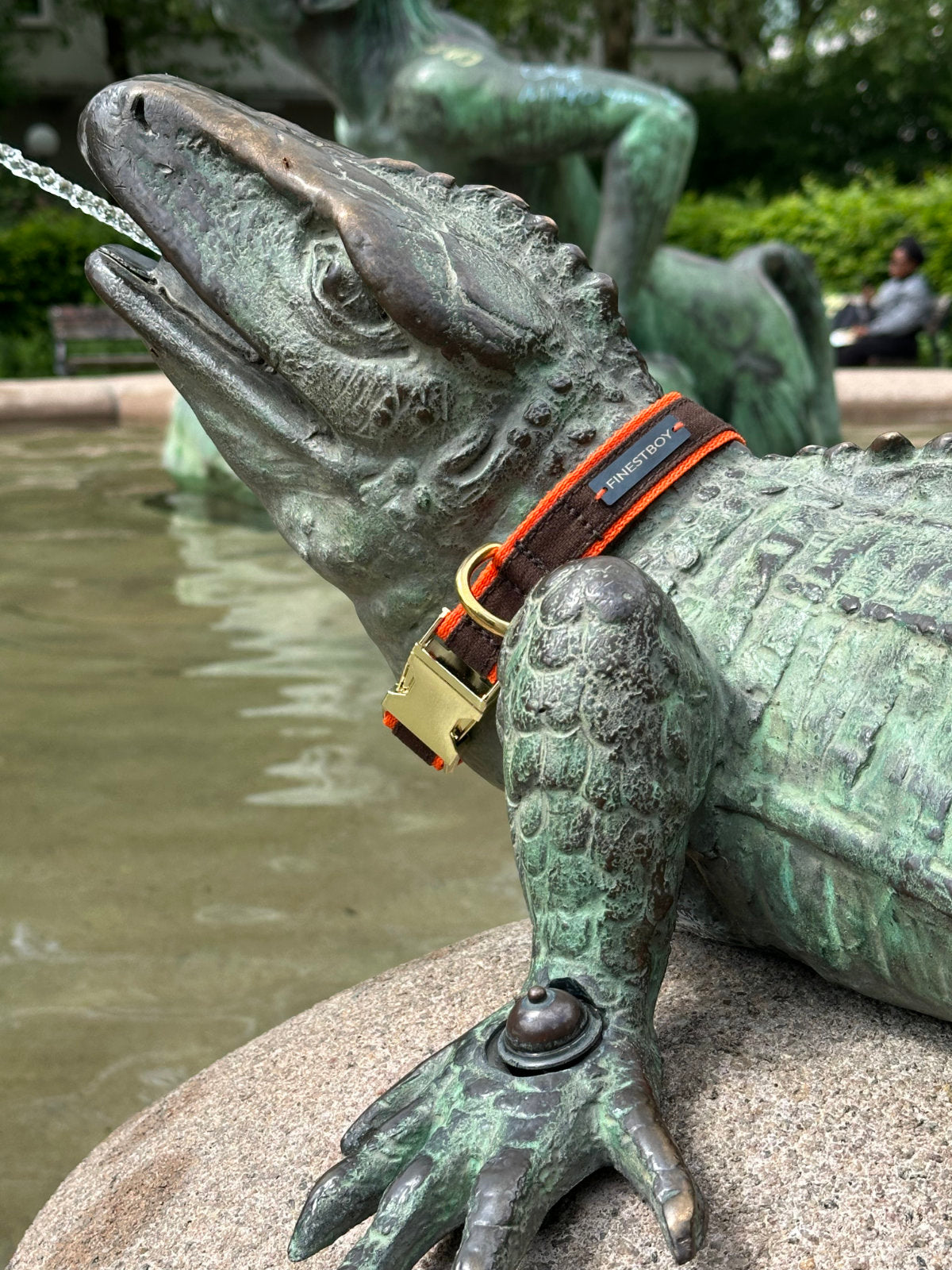Handgefertigtes FINESTBOY-Halsband, 25mm, orangefarbenes Gurtband mit braunem Canvas-Streifen und Messing-Applikationen, Nahaufnahme an Echsen-Skulptur am Brunnen in Hamburg-Altona.