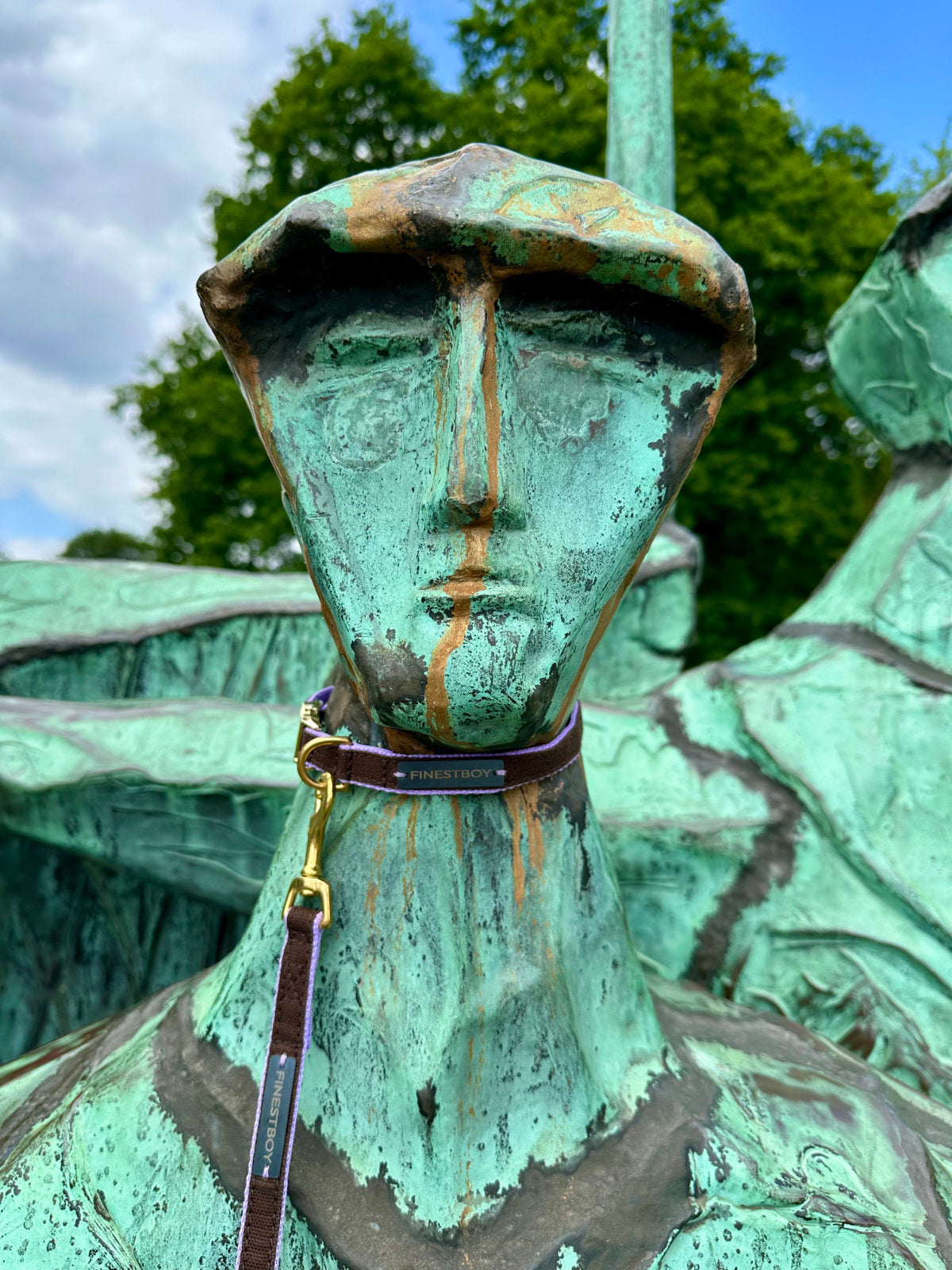 Metallskulptur am Hamburger Altonaer-Balkon, trägt FINESTBOY-Bundle aus Halsband und Leine, 15mm, fliederfarbenes Gurtband mit dunkelbraunem Canvas-Streifen und Vollmessing-Applikationen, vermessingter Metallsteckschnalle und Vollmessing-Karabiner.