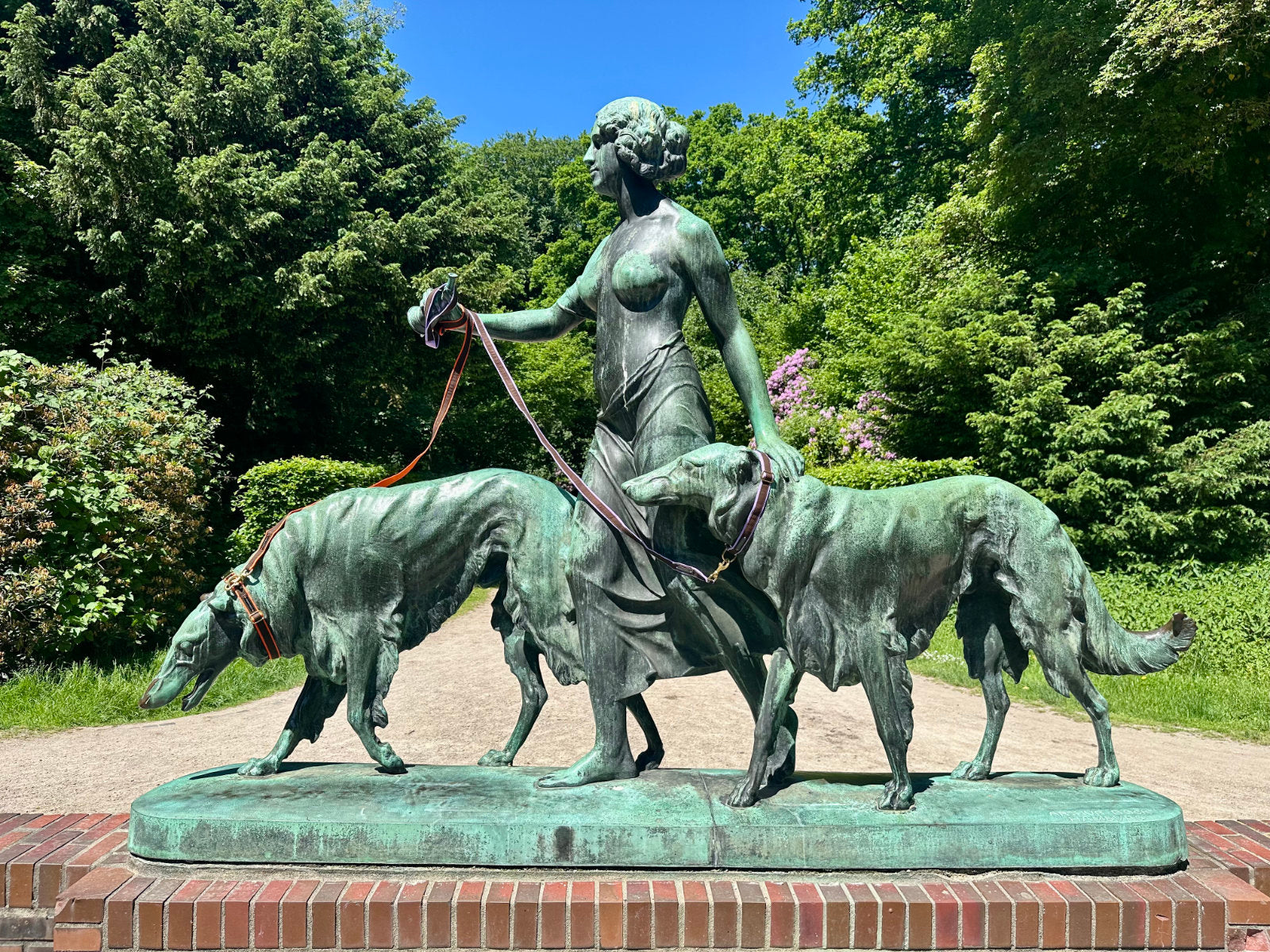 Bronzestatue einer Frau mit zwei Windhunden im Hamburger Stadtpark, die Hunde tragen handgefertigte FINESTBOY-Bundles aus Halsband und Leine, 25mm, Gurtband in Orange und Flieder mit dunkelbraunem Canvas und Vollmessing-Applikationen und vermessingten Metallsteckschnallen.