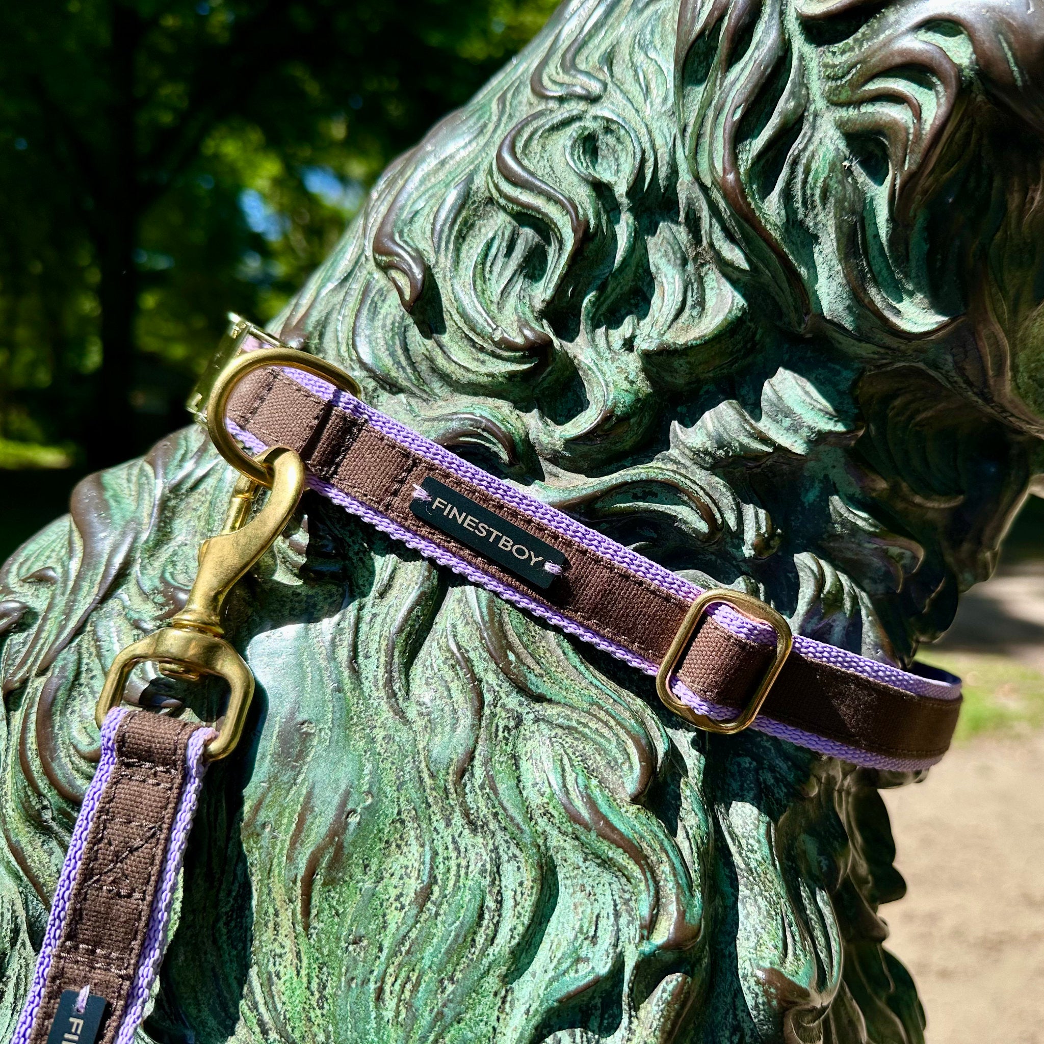 Hund trägt FINESTBOY-Bundle aus Halsband und Leine, 25mm, fliederfarbenes Gurtband mit dunkelbraunem Canvas-Streifen und Vollmessing-Applikationen, vermessingter Metallsteckschnalle und Vollmessing-Karabiner, im Hamburger Stadtpark an einer Hundeskulptur