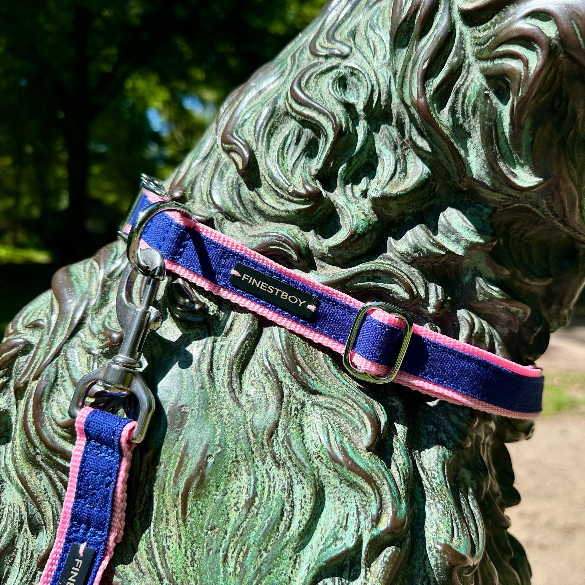Hund trägt FINESTBOY-Bundle aus Halsband und Leine, 25mm, rosafarbenes Gurtband mit royalblauem Canvas-Streifen und verchromten Vollmessing-Applikationen, verchromter Metallsteckschnalle und Edelstahl-Karabiner, im Hamburger Stadtpark an einer Hundeskulptur