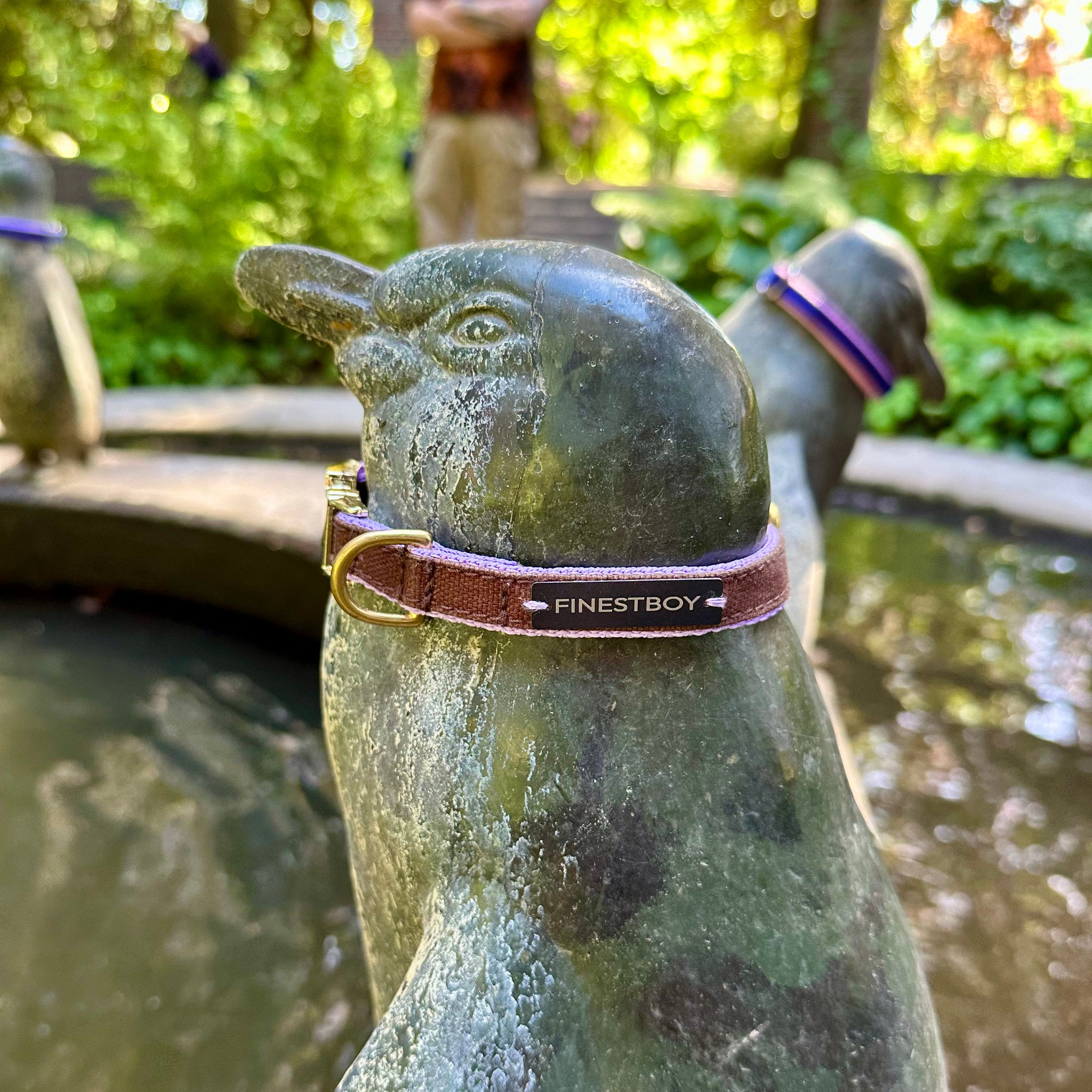 Pinguin trägt FINESTBOY-Halsband, 15mm, fliederfarbenes Gurtband mit dunkelbraunem Canvas-Streifen und Vollmessing-Applikationen und vermessingter Metallsteckschnalle, im Hamburger Stadtpark an einer Pinguin-Skulptur