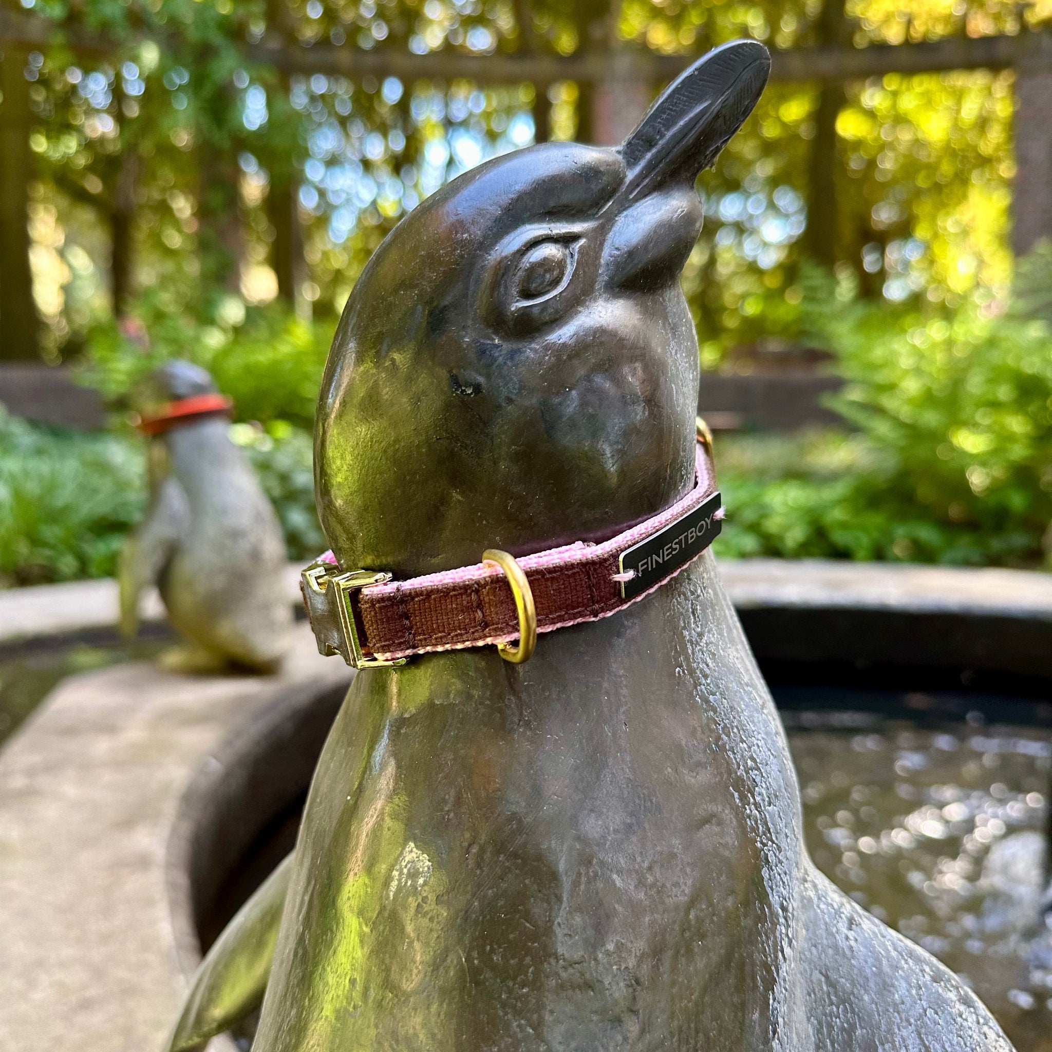 Pinguin trägt FINESTBOY-Halsband, 15mm, rosafarbenes Gurtband mit dunkelbraunem Canvas-Streifen und Vollmessing-Applikationen und vermessingter Metallsteckschnalle, im Hamburger Stadtpark an einer Pinguin-Skulptur