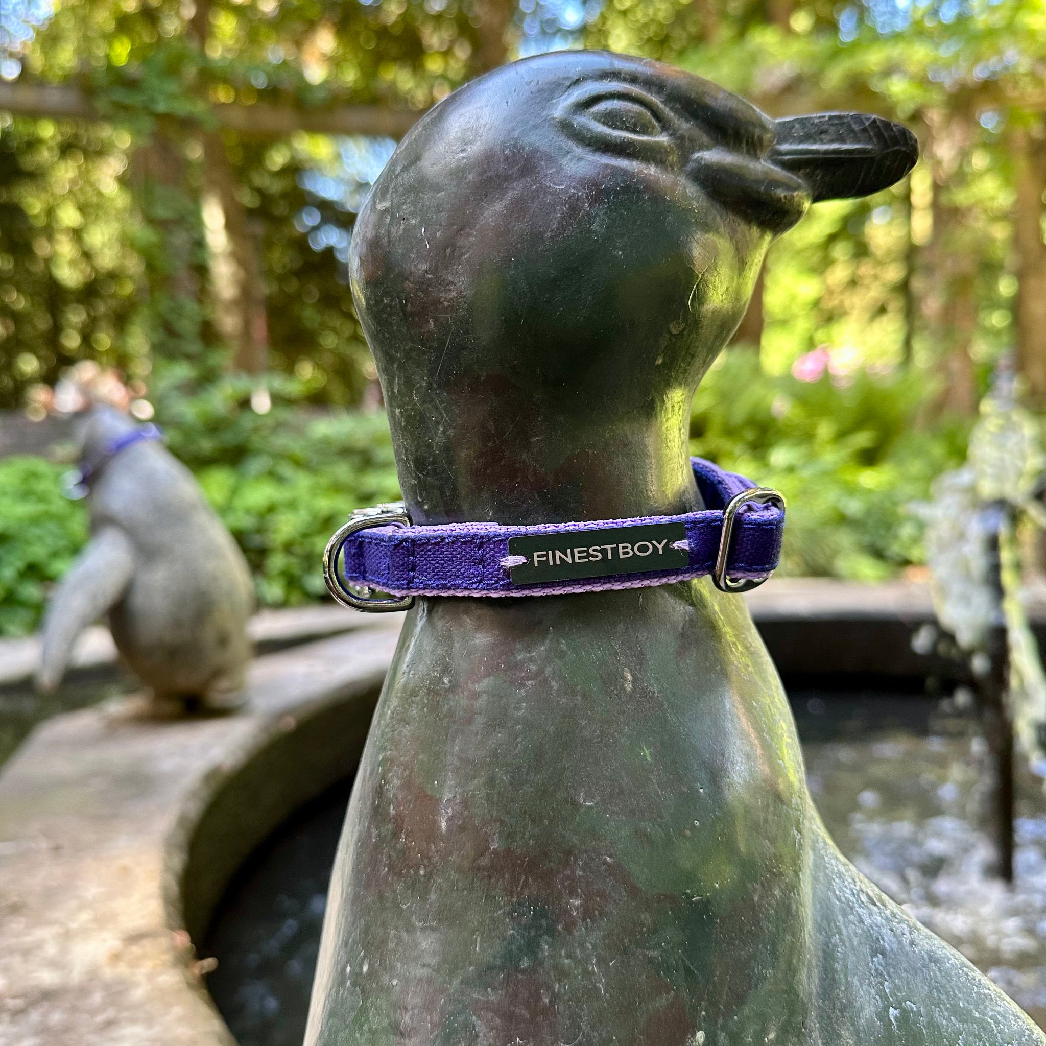 Pinguin trägt FINESTBOY-Halsband, 15mm, fliederfarbenes Gurtband mit royalblauem Canvas-Streifen und verchromten Vollmessing-Applikationen und verchromter Metallsteckschnalle, im Hamburger Stadtpark an einer Pinguin-Skulptur